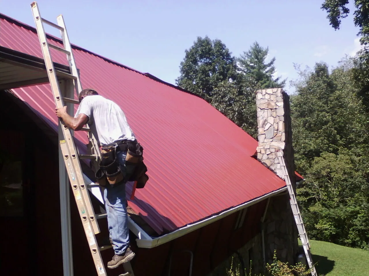 Roofer on a ladder beside a red metal roof and stone chimney — Soffit & Fascia Repair by Roofing Brothers in Griffin